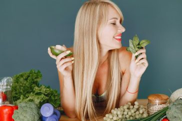 A smiling woman holding avocado and basil, surrounded by fresh vegetables and fruits for healthy eating.