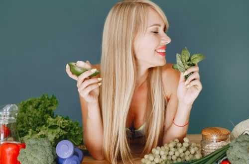 A smiling woman holding avocado and basil, surrounded by fresh vegetables and fruits for healthy eating.
