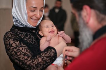 A smiling baby being baptized in a church, held by a woman with a headscarf.