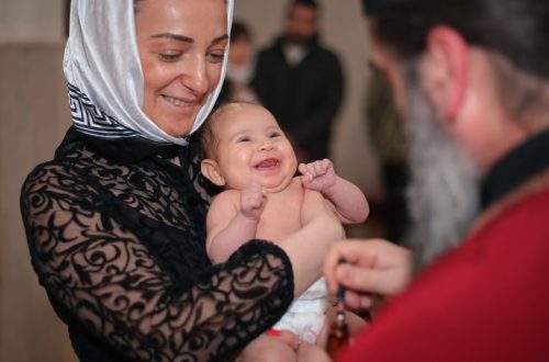 A smiling baby being baptized in a church, held by a woman with a headscarf.